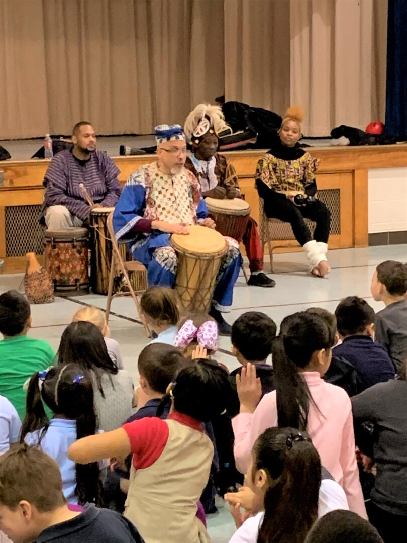 Children sitting on the floor watching four people play hand drums in front of a stage