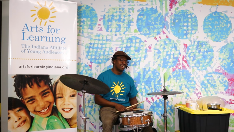Man smiling sitting at a drum set with an Arts for Learning banner next to him