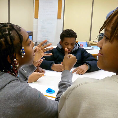 Photo of black girl talking to a group of students with girl to the tight of her smiling and boy in front of her looking at paperwork